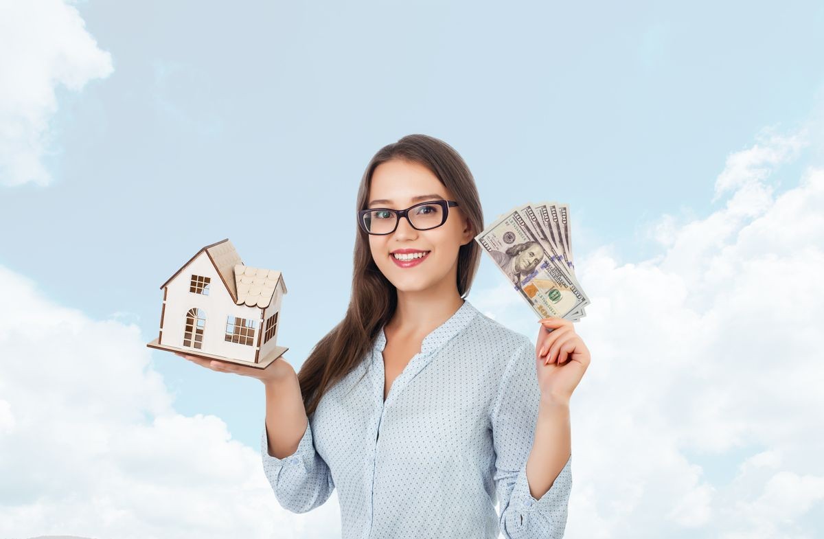Smiling beautiful girl holding house and dollar banknotes on background of sky. Building, mortgage, real estate, sale, finance and property concept.